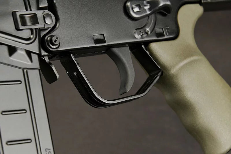 Close-up of a rifle receiver area with a black trigger guard and a tan/greenish grip against a dark gray background.