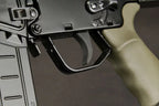 Close-up of a rifle receiver area with a black trigger guard and a tan/greenish grip against a dark gray background.