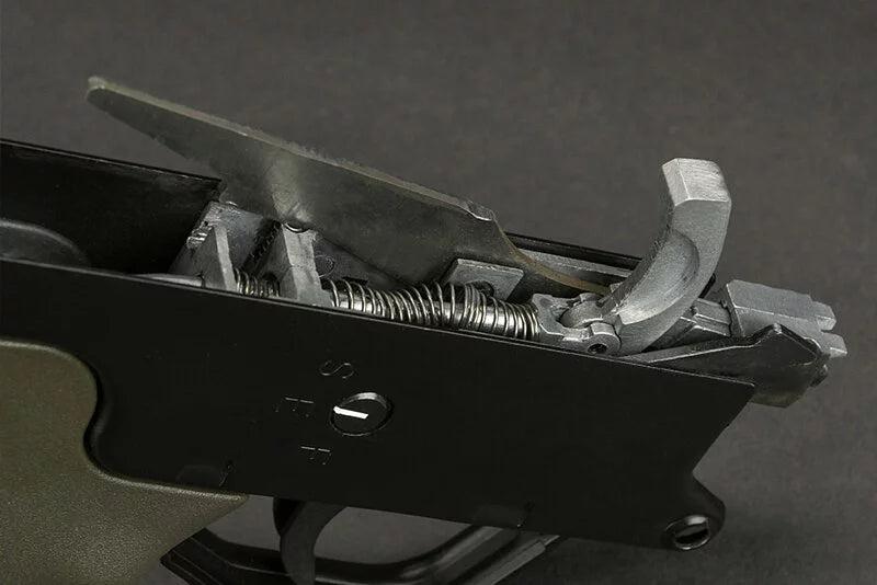 Close-up of a firearm's trigger mechanism showing the black receiver, silver metal spring, and trigger components against a dark gray background.