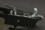 Close-up of a firearm's trigger mechanism showing the black receiver, silver metal spring, and trigger components against a dark gray background.