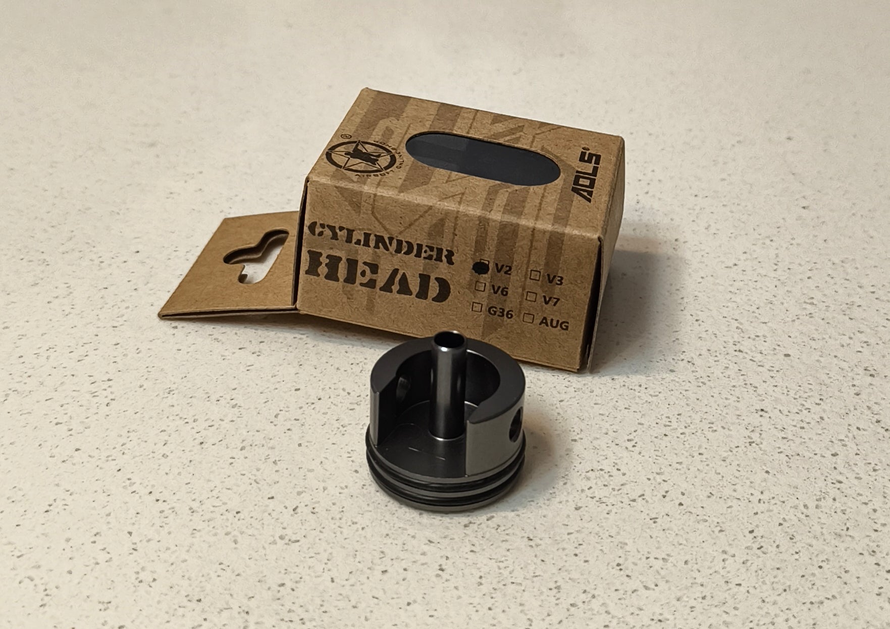 Black cylinder head on a light gray surface with a cardboard box labeled 'Cylinder Head' in the background.